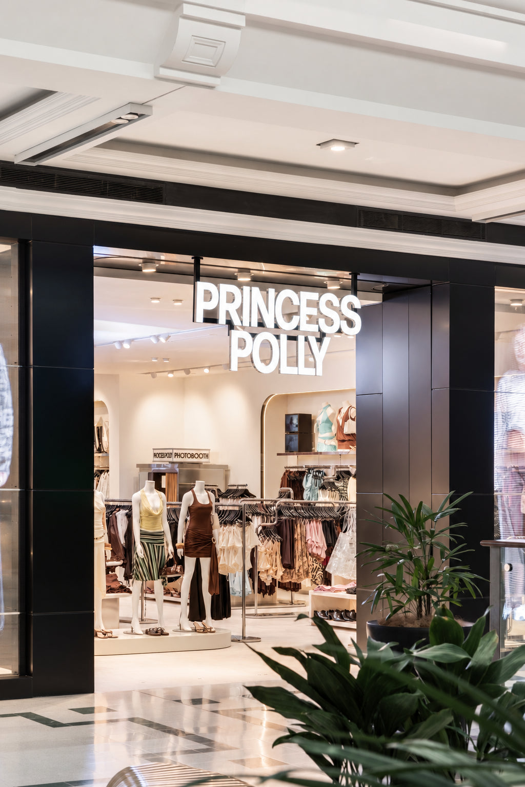Princess Polly clothing store entrance with illuminated white brand signage above a wide opening, showing mannequins and racks of apparel inside.